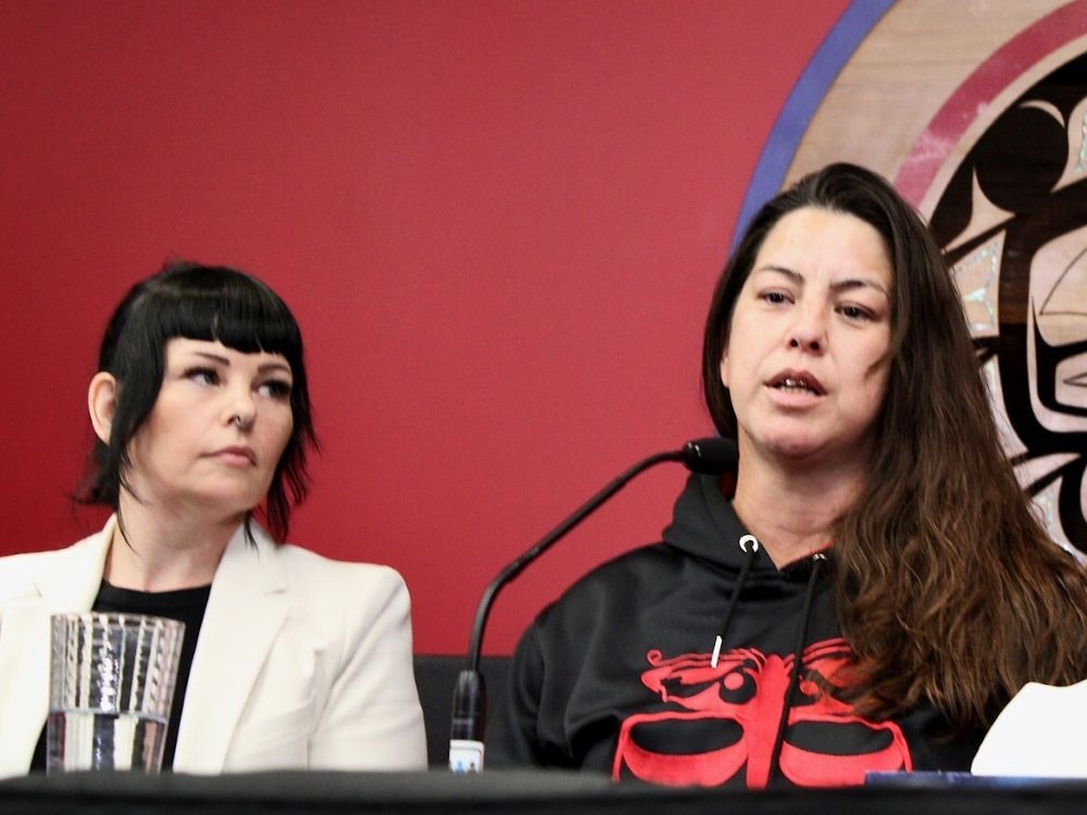 Two women sit at a table with microphones. The woman on the left wears a white suit and has black hair with bangs. The woman on the right has long brown hair and wears a black and red sweatshirt.
