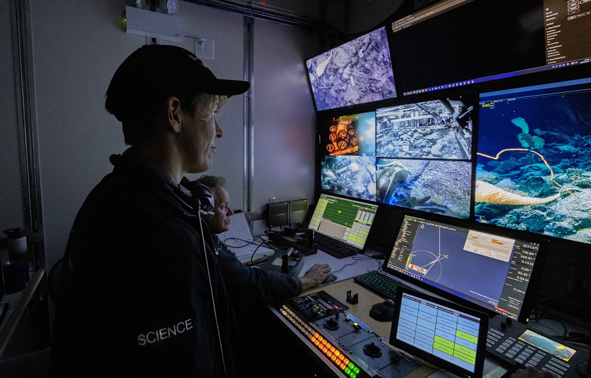 A woman with short blond hair covered by a baseball cap stands in front of a wall of screens that show what a remotely operated vehicle can see. 