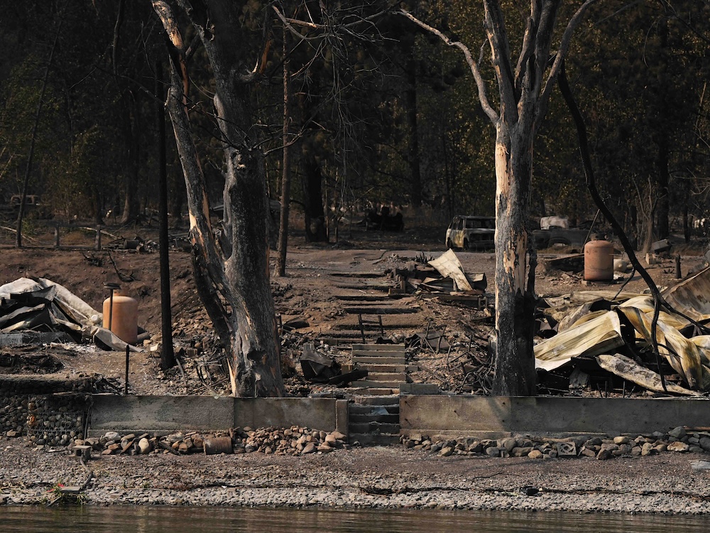 Burned trees and rubble from two burned structures can be seen on the shore of a lake.