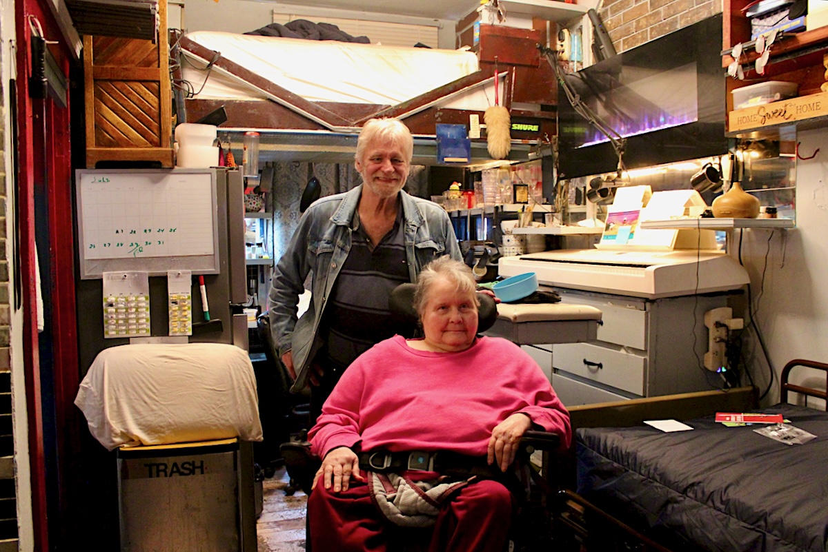 A man in his 70s stands behind a small woman also in her 70s who uses a wheelchair. The people are inside a small room that is well organized but crammed with furniture.