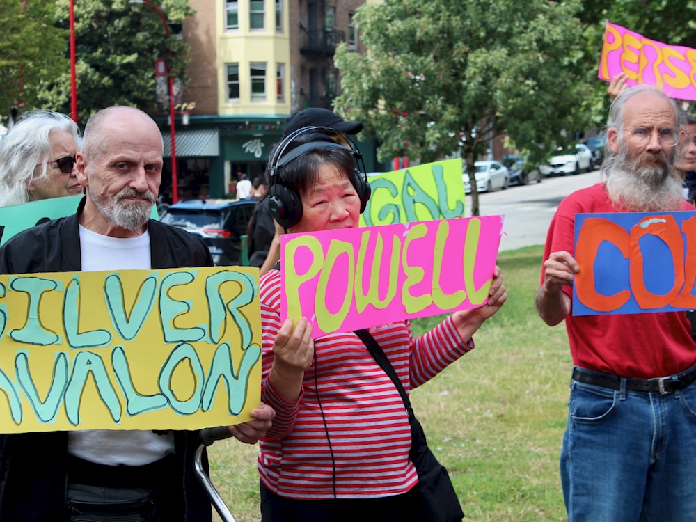 Two elderly men and an older woman stand in a city park holding signs that say the names of SRO hotels, including the Powell and the Silver Avalon.