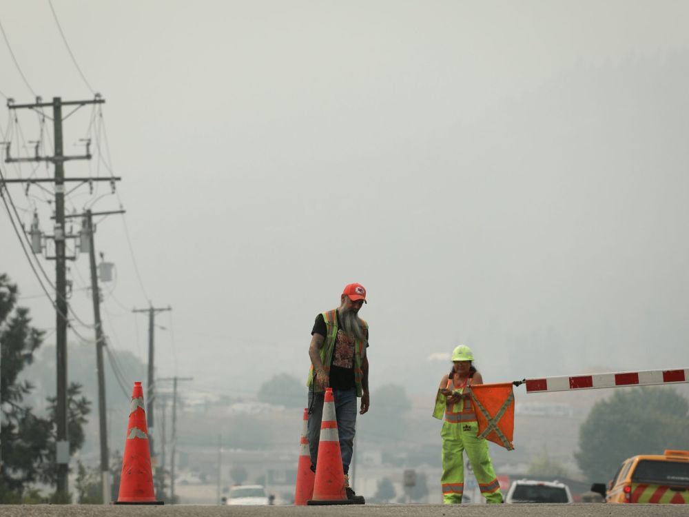 Workers in hard hats and high-viz safety vests control vehicle traffic on a highway against a background of gray, smoky skies.