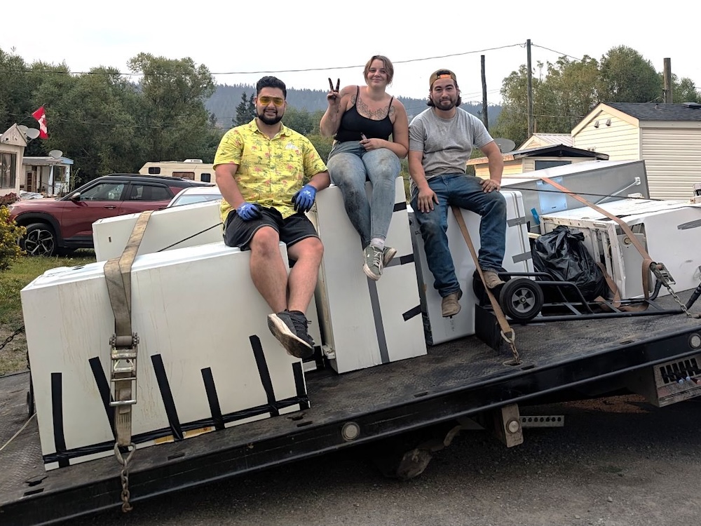 Three young people sit smiling on used freezers on a flat-deck trailer.