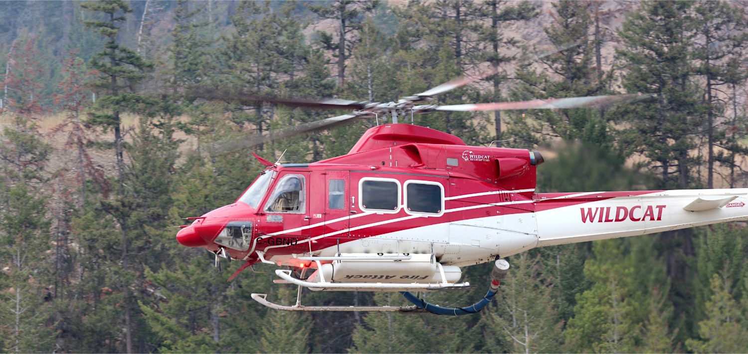 A large red helicopter with a contraption on the bottom and a large visible hose flies in front of an arid forested mountainside.