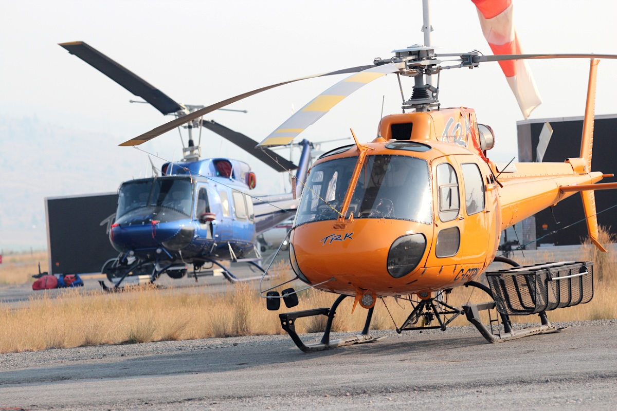 Two helicopters sit on a helipad at a smoky base, with yellow grass visible.