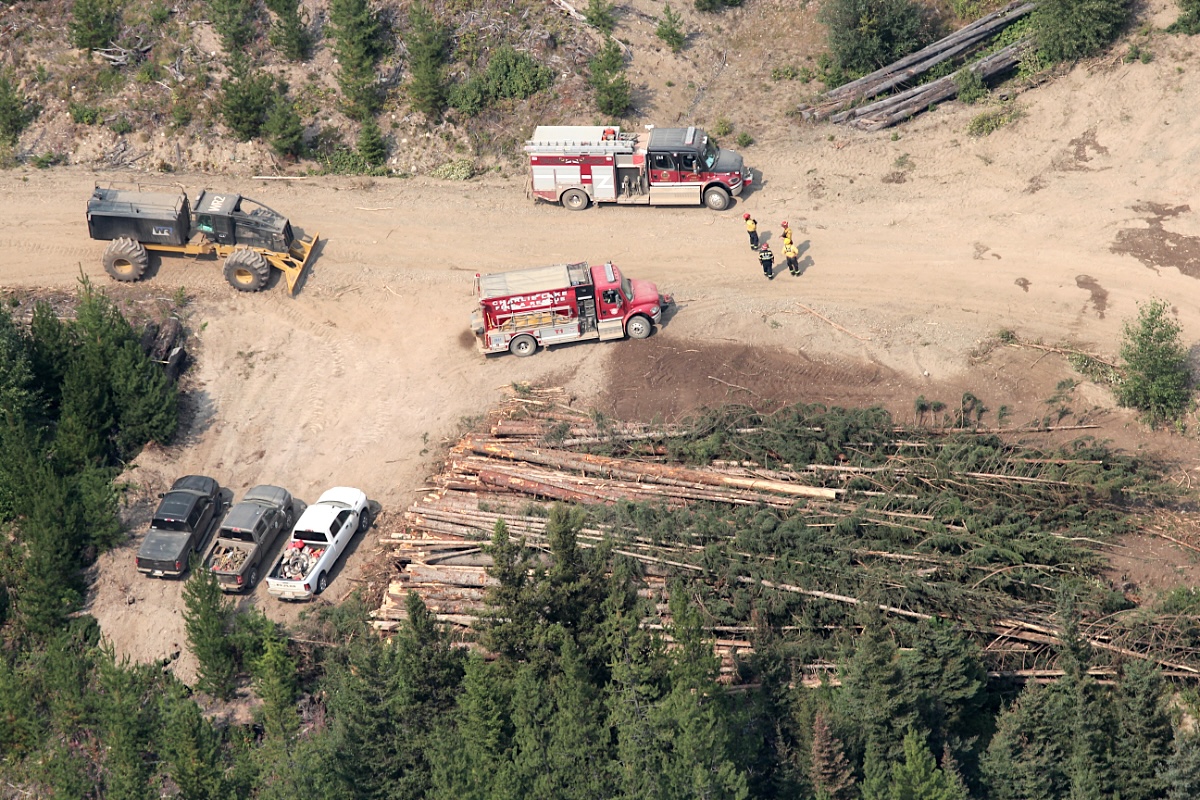 A bulldozer, pickup trucks, log piles, fire trucks and four firefighters are visible from above on a remote forestry road. 