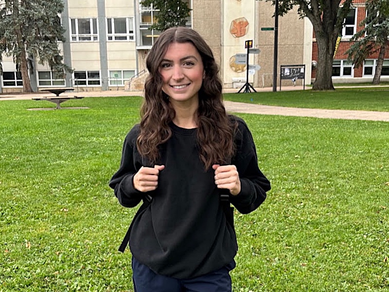 A young woman with light skin tone and long brown hair stands on a lawn in front of a school building.