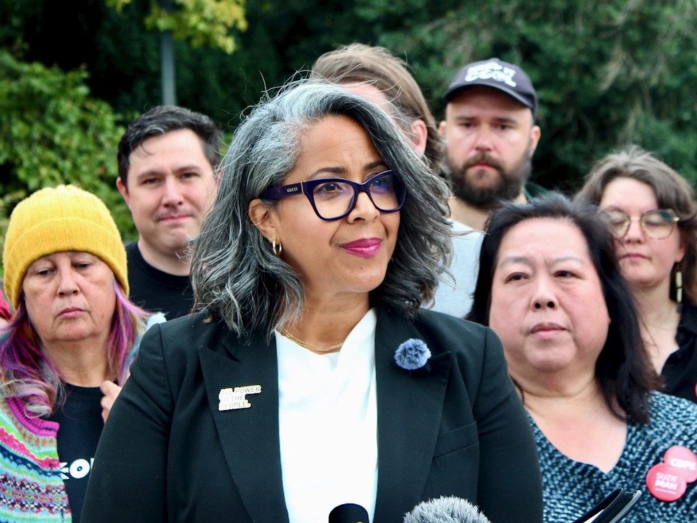 A woman with grey and black shoulder-length hair and medium skin tone stands in front of broadcast news microphones.