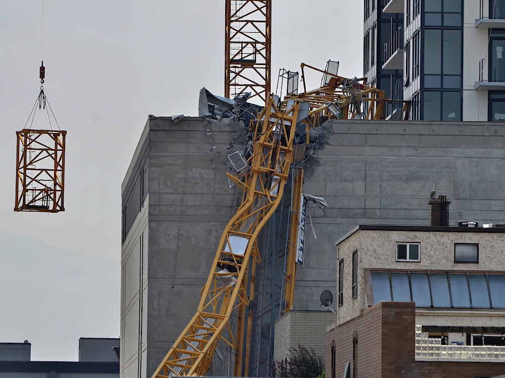 A portion of a yellow metal construction crane lies mangled against the side of a building in a downtown area.