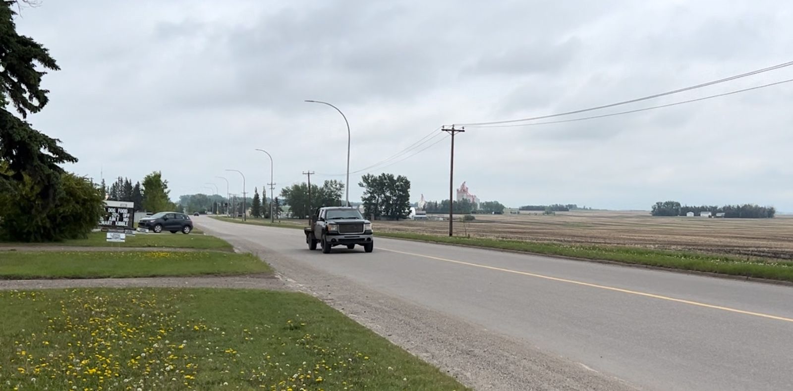 An empty field lies across a road. On the other side are houses and yards. A truck drives down the road.