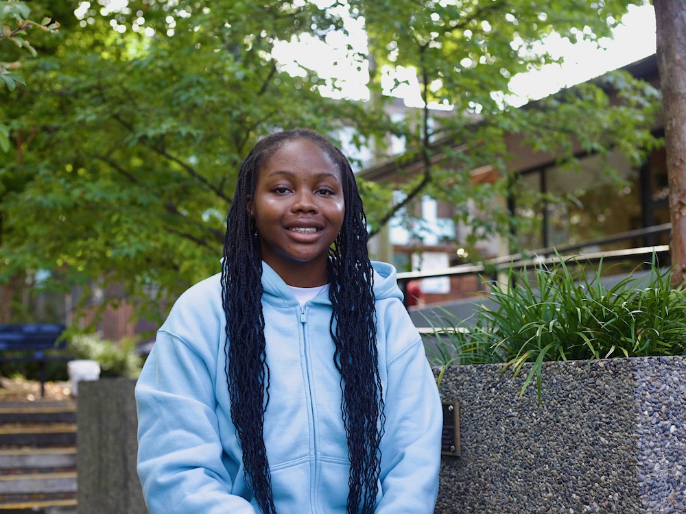 A young woman with long black hair and dark skin smiles at the camera. She sits in an outdoor area and wears a light blue zip-up hoodie.