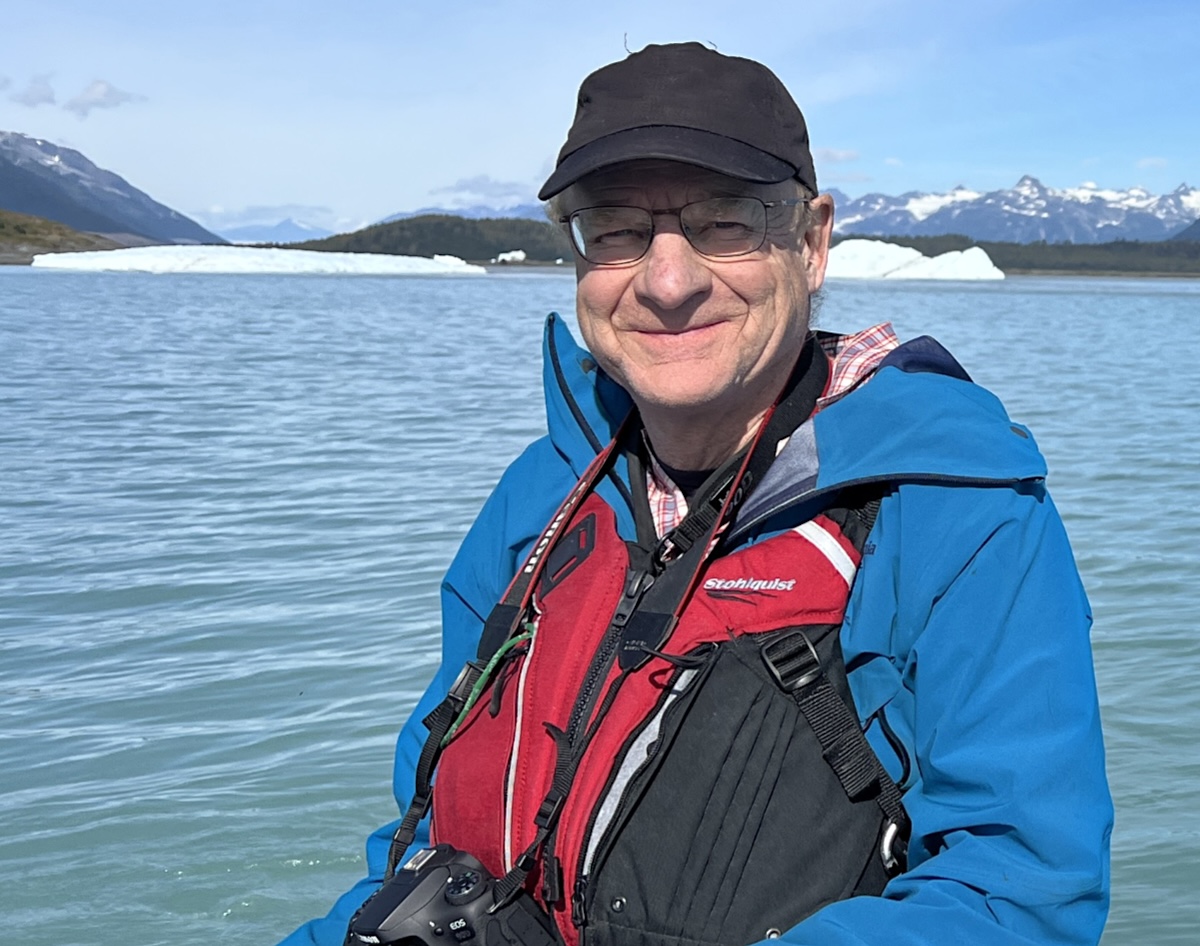 A man with light skin tone and wearing a life preserver and black ball cap smiles while sitting on a boat in a glacial lake. Calving glaciers are visible behind him.