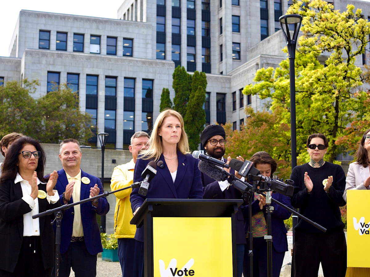 A woman with long blond hair stands at a lectern with people surrounding her and Vancouver city hall in the background.