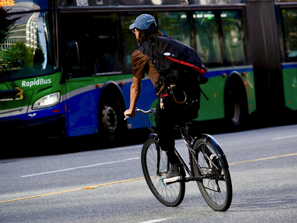 A man wearing a brown shirt, a backpack and a cap can be seen from behind as he rides a bicycle on a city street. A bus can be seen in the background.