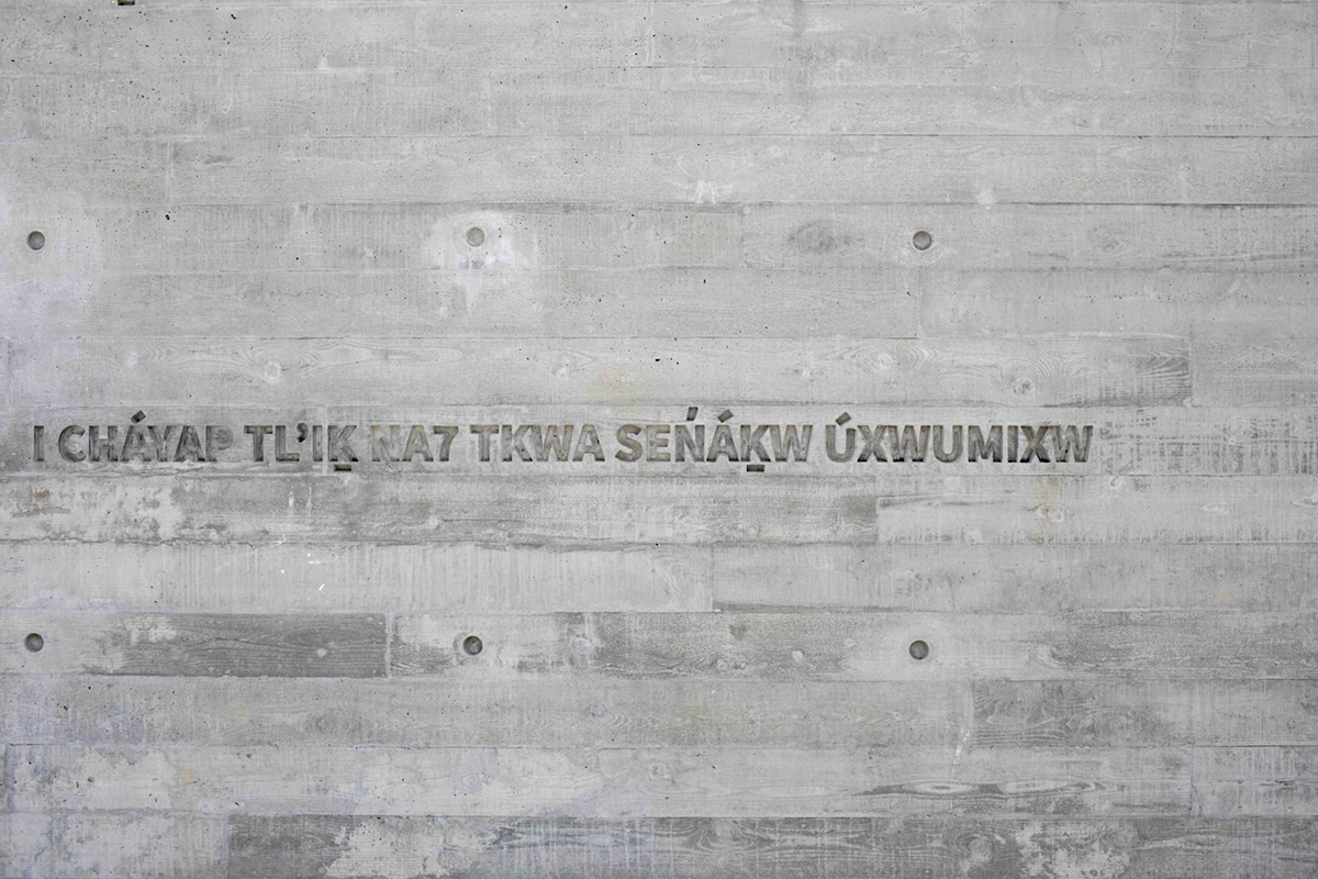 A grey plank of wood is etched with bold sans-serif typeface in the Sḵwx̱w&uacute;7mesh language. The phrase translates to &lsquo;You have arrived at Sen̓&aacute;ḵw.&rsquo;
