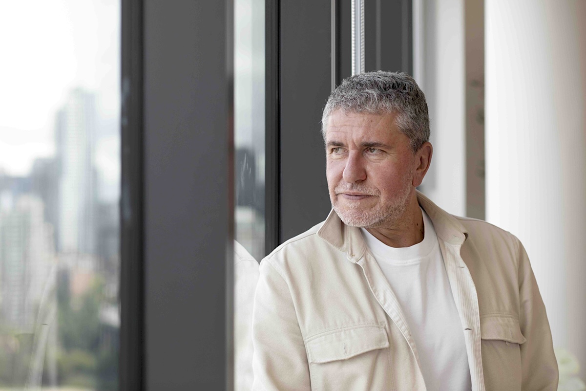 Venelin Kokalov is wearing a cream-coloured chore coat over a white T-shirt. He has short grey hair and a light beard. He leans against a window, looking to the left of the frame.