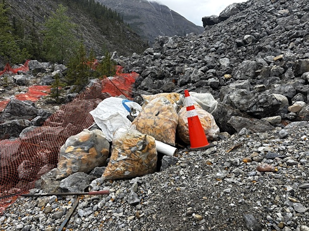 Multiple garbage bags lie beside toppled-over orange fencing off the Alaska Highway. There&rsquo;s an orange traffic cone and a sea of rocks beside the cleanup site. 