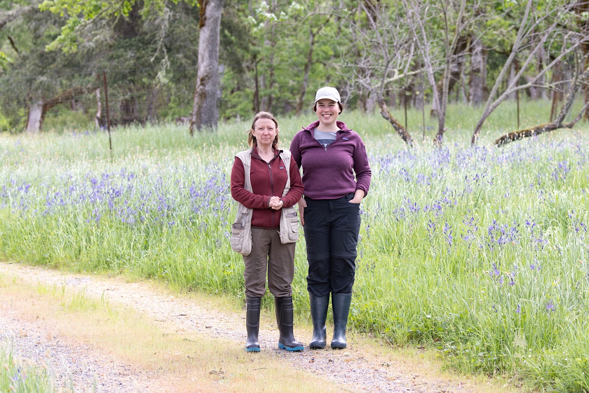 Two women stand beside a camas meadow. They are wearing warm clothes and rubber boots.