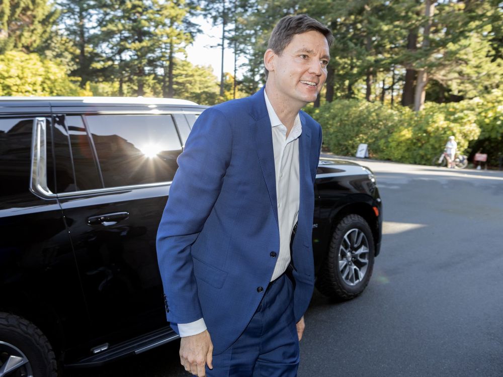 A tall light-skinned man in a blue suit with no tie smiles as he steps out of a large black vehicle.