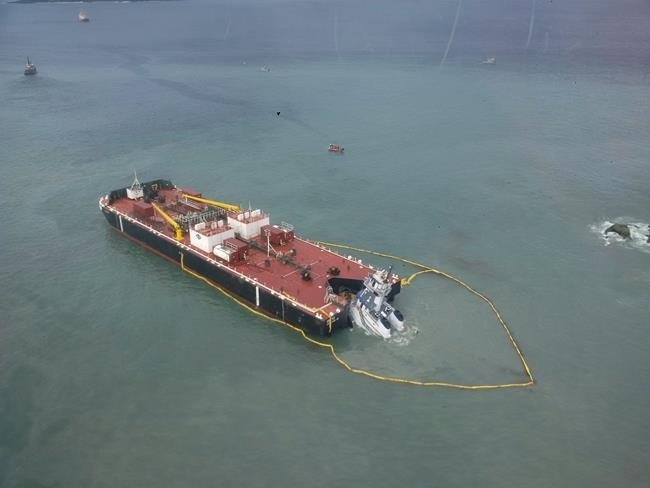 An aerial photo of a large barge with a red deck and a sunk tug at its rear, with floating yellow barriers to contain spilled diesel fuel.