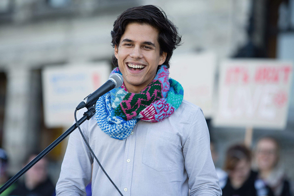 A young man in his late teens or early 20s with dark brown hair and medium-light skin and wearing a scarf speaks at a microphone.
