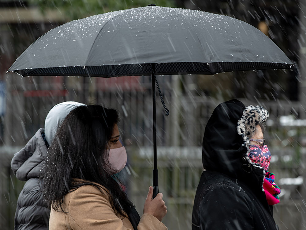 Three people stand under a black umbrella in the snow. They are facing the right of the screen. They are wearing masks.