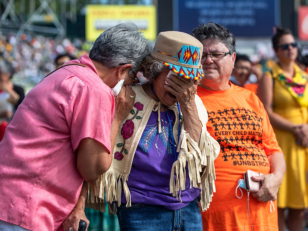An elderly woman in a crowd is supported on either side by friends as she bends her head weeping.