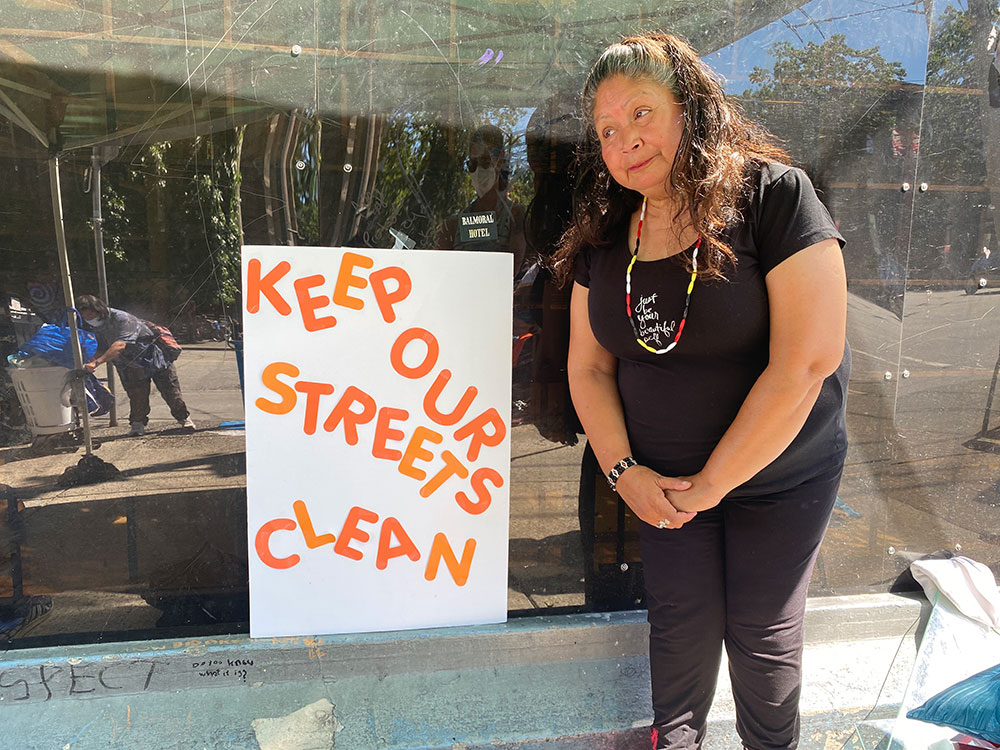 An Indigenous woman, Mama Marcy, is standing to the right of a handmade sign, white posterboard with orange letters, that reads &ldquo;Keep our streets clean.&rdquo; Marcy is wearing black and a red, black and white beaded necklace in the colours of the medicine wheel.