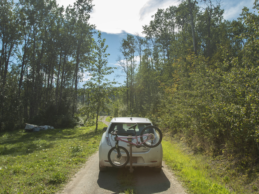 A Nissan Leaf on a leafy rural lane. A bicycle is affixed to the bike rack.