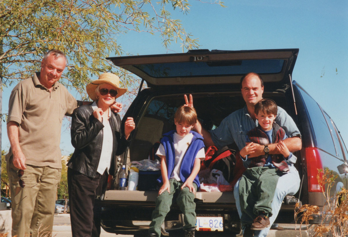 Five people stand in front of the open back of a sports utility vehicle on a sunny day. Glen Fawkes is wearing a blue shirt and holding one of his sons in his lap. He poses jokingly for the camera, holding a peace sign behind his son Kyle&rsquo;s head. Kyle is wearing a blue vest.
