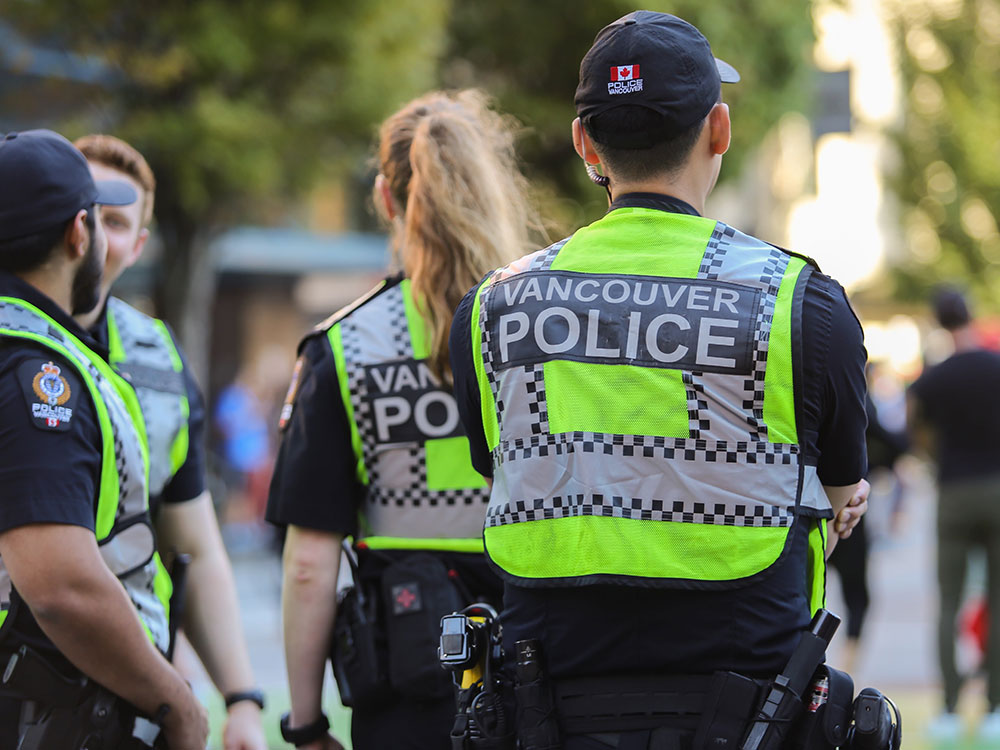 Four Vancouver police officers stand with their backs to the camera. They are wearing fluorescent yellow vests over their uniforms that read “Vancouver Police." The sunny, green background is out of focus; the officers are at a September 2022 protest decrying the murder of Masha Amini.