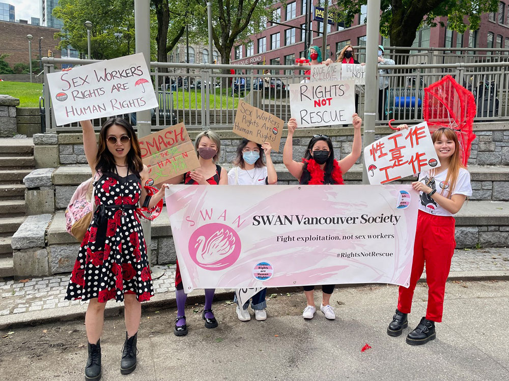 Five people stand at Victory Park in Vancouver. One person holds a red umbrella. Everyone holds signs reading things such as, &ldquo;Sex Workers Rights Are Human Rights&rdquo; and &ldquo;Rights not Rescue.&rdquo; Two signs feature Chinese characters. 