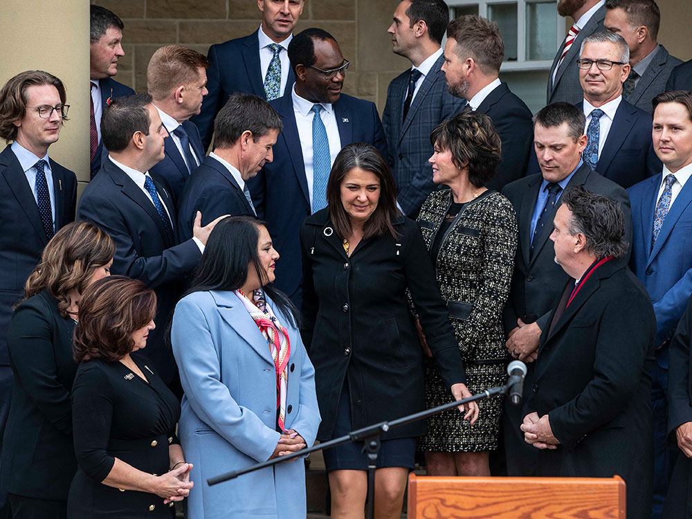 A crowd of people fills the frame. They are wearing dark suits &mdash; navy and black &mdash; and standing on a flight of outdoor stairs. Alberta premier Danielle Smith makes her way towards the podium at the bottom of the frame. She has dark shoulder-length hair and is wearing a black jacket and navy skirt. The people around her, her new cabinet members, are turned towards her and making space for her to descend the stairs.