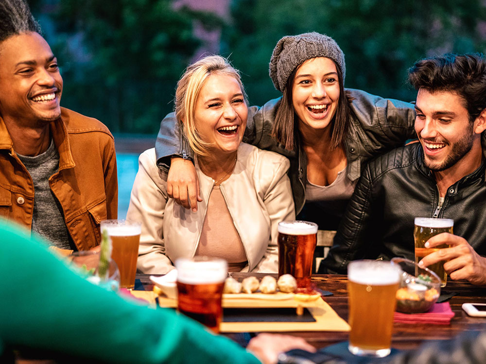 A group of smiling friends gathers around a table on an outdoor patio over pints of beer and plates of appetizers. At the centre of the frame is a woman in a grey toque and black leather jacket. She is looking up and smiling, and putting her arms around two friends: a woman with blonde hair in a white to the left, and a man with dark hair and a black jet to the right.