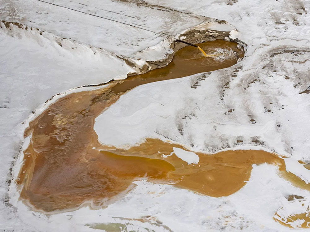 An aerial photo shows a snowy, treeless. A pipe pours brown water into a pond, and the overflow stretches over a wide area.