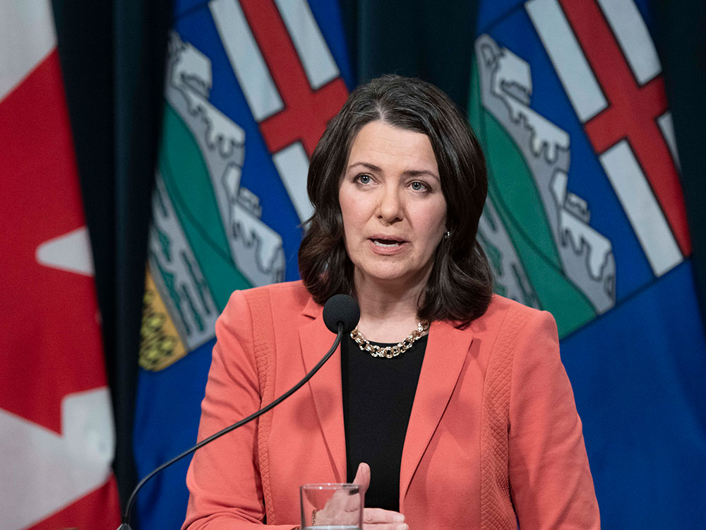A woman in an orange blazer, Premier Danielle Smith, stands at a podium, emblazoned with the word ‘Alberta,’ and answers reporters’ questions during a news conference in Calgary on Tuesday, Jan. 10, 2023. Albertan and Canadian flags are draped behind her.