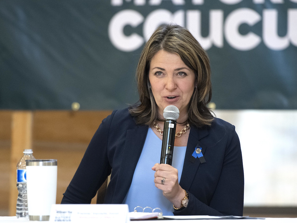 Danielle Smith, wearing a baby blue blouse and navy blazer, holds a microphone at a podium while speaking.