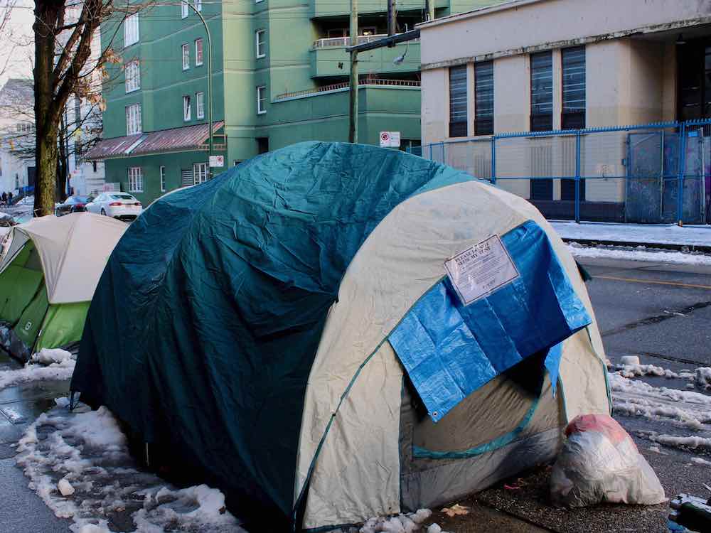 A tent with a dark teal cover is in the foreground, with a smaller green tent behind it. The tents are on a sidewalk in Vancouver&rsquo;s Downtown Eastside. There is snow on the ground and the sky is blue.