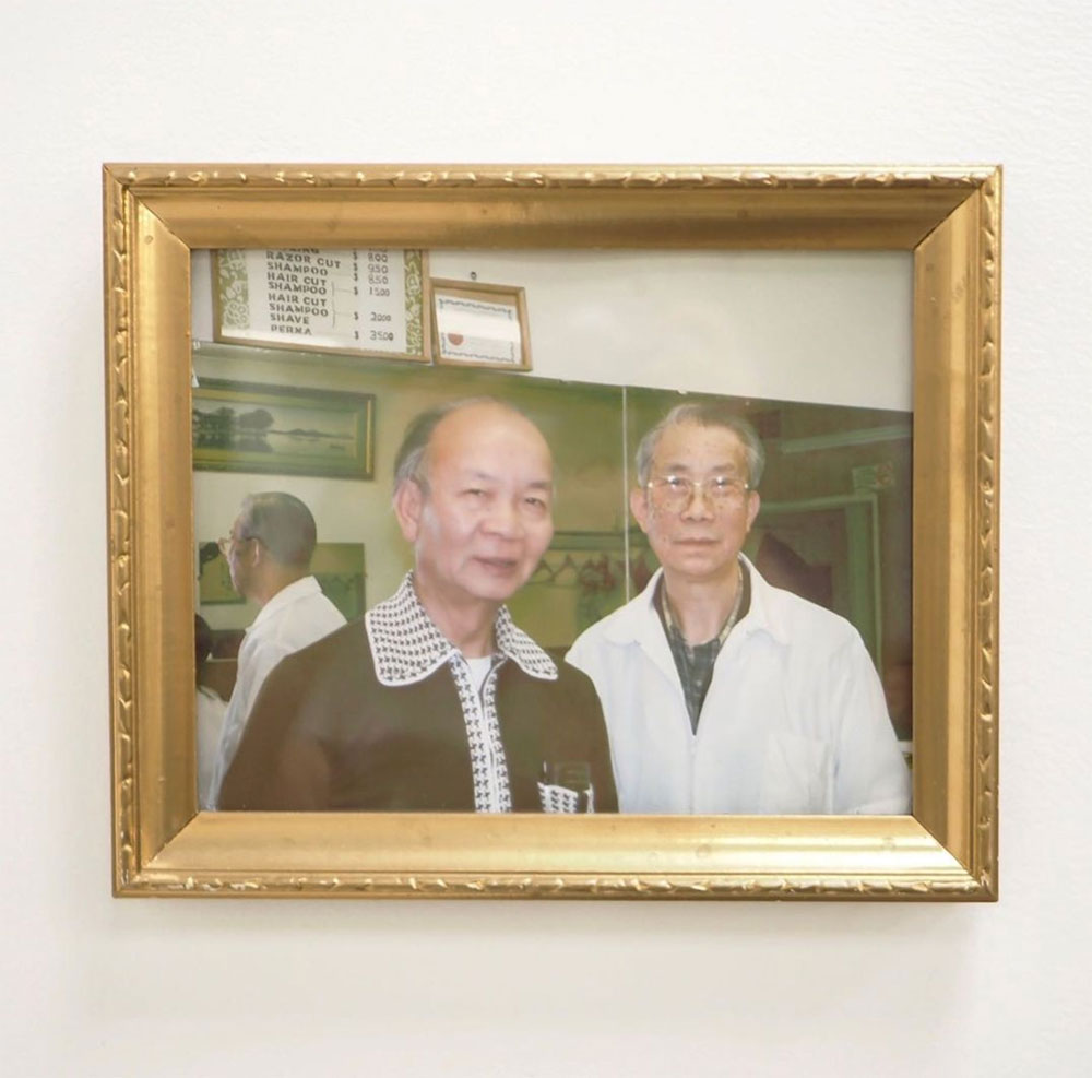An old colour photo in a gold frame hangs on a white wall. It depicts Jack Gee, left, an elderly Chinese man in a brown cardigan, standing in a barber shop next to his friend Roy, right, in a white barber&rsquo;s coat over a plaid shirt.