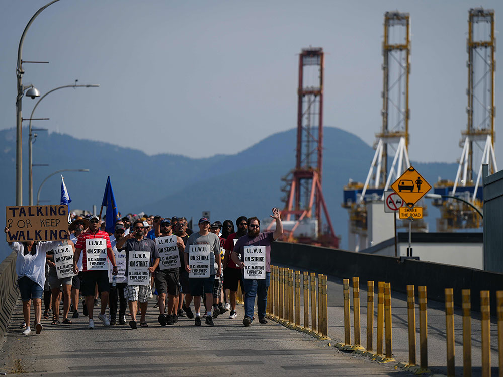 A large number of people carrying protest signs walk across a bridge toward the camera, with the Port of Vancouver&rsquo;s giant cranes in the background.