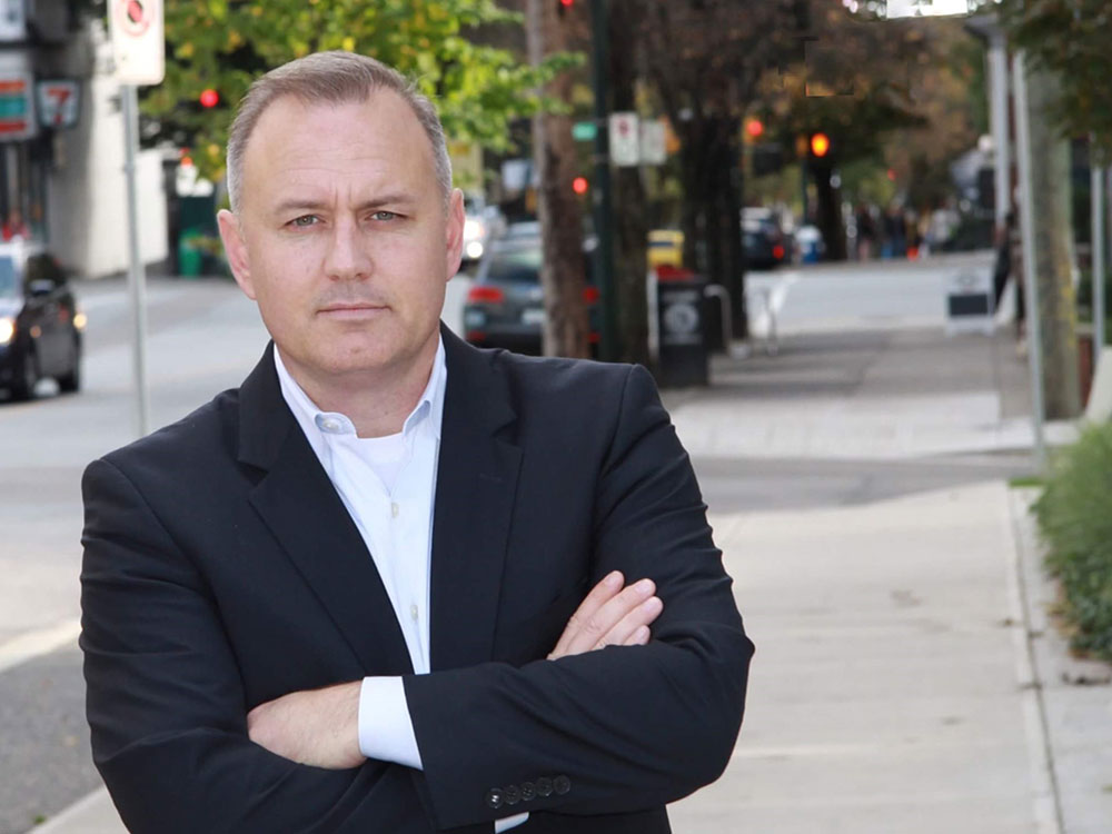 A white middle-aged man with silver hair stands on a sidewalk in a suit jacket and open collared white shirt with arms folded, looking intently at camera.