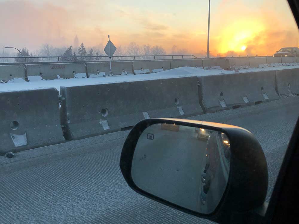 The view from the driver&rsquo;s seat of a car at sunrise on a winter day. Concrete barricades are covered in a light dusting of snow. On the horizon, the grey sky is turning orange.