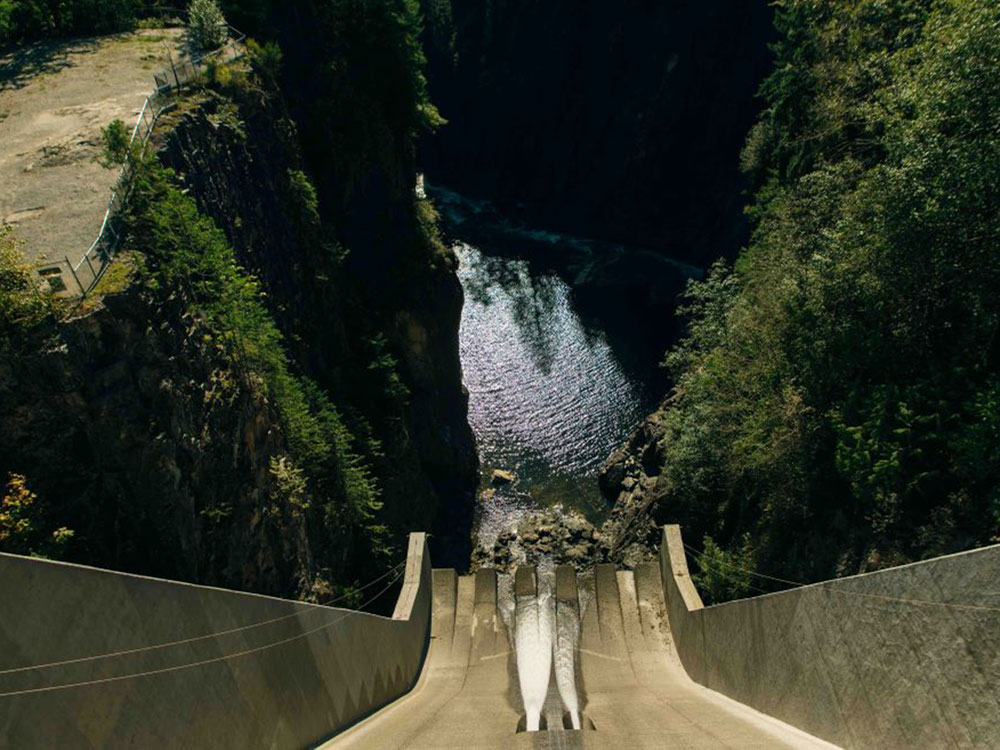 The view from the top of the Cleveland Dam, a tall concrete structure that divides the Capilano River below from the water reservoir out of sight. At the bottom of the concrete, two white jets of water lead to a rocky riverbed. The river is flanked by stands of tall coniferous trees.