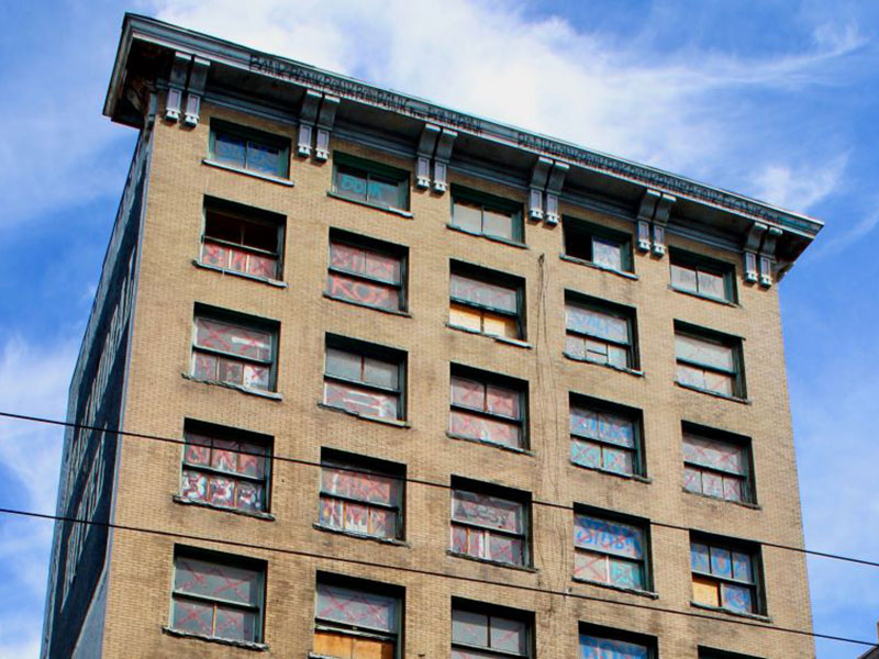 A tall brick building stands against a bright blue sky. Many of the windows are boarded up and are tagged with graffiti.