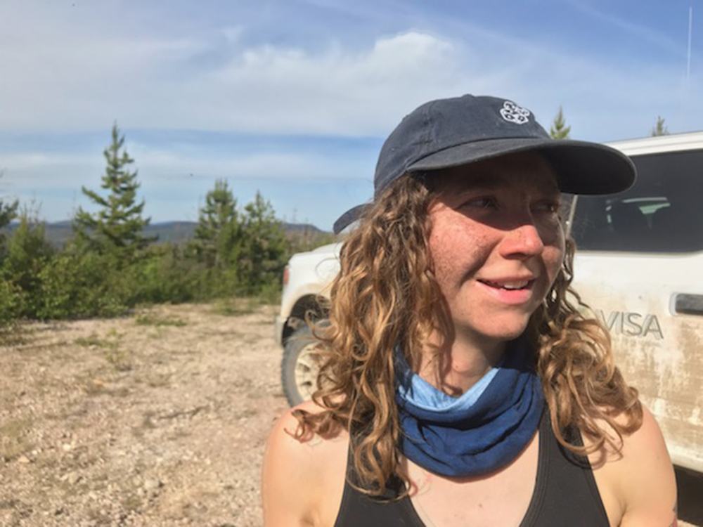 A woman with curly hair and freckles, wearing a blue hat and bandana, stands in front of a white truck on a cut block.