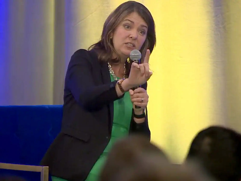 A white woman with shoulder-length brown hair, wearing a blue jacket, green shirt and heavy necklace, sits on a chair holding a microphone and holding up one hand.
