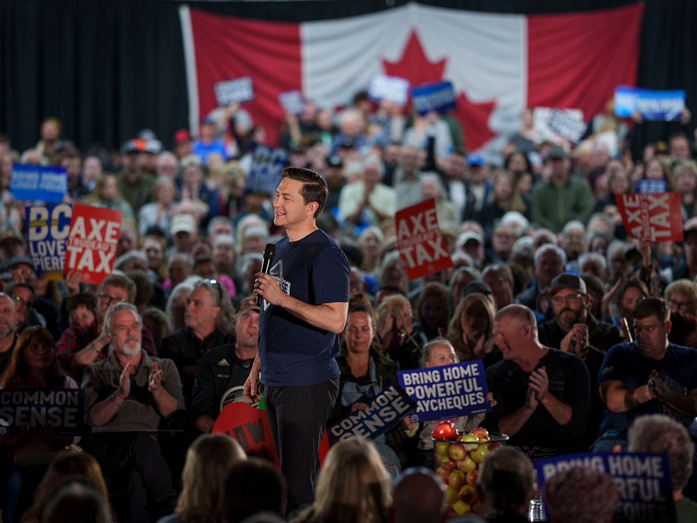 A white man with short hair, wearing a blue T-shirt and holding a microphone, stands amid a large audience in a darkened hall.
