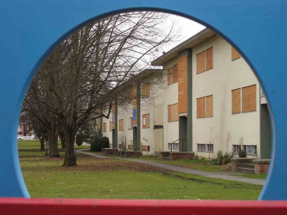 Framed by a blue circular border part of a children&rsquo;s play structure, a row of beige three-storey walk-up apartment buildings stands along a tree-lined street. Their windows are boarded up with plywood.