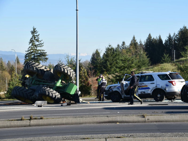A large green tractor is laying on its side on the highway, wheels pointing to the sky. Police cars, one damaged on its side, are parked nearby.