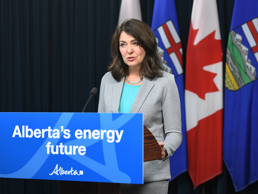 A 52-year-old white woman with shoulder-length dark brown hair, wearing a turquoise shirt and pale grey suit, stands at a podium with a sign saying 'Alberta’s energy future.' Behind her are Albertan and Canadian flags.